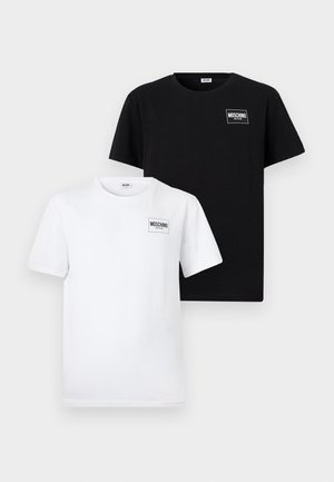 Homme aux cheveux bouclés et foncés, portant un t-shirt noir et un boxer noir, debout pieds nus devant un fond clair uni.