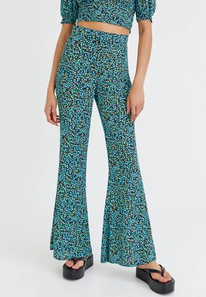 Spodnie z wysokim stanem i rozszerzanymi nogawkami w niebieski, zielony i biały wzór kwiatowy, noszone z czarnymi sandałami na platformie.