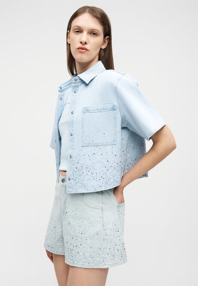 Νεαρή γυναίκα που φοράει ανοιχτό μπλε πουκάμισο από τζιν με κοντά μανίκια και κουμπιά, καθώς και ταιριαστό σορτς με διάσπαρτο σχέδιο από μαύρες πιτσιλιές βαφής.