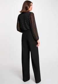 Blouse noire avec des manches bouffantes transparentes et des pois argentés ; pantalon noir taille haute avec une coupe ajustée et des poches latérales.
