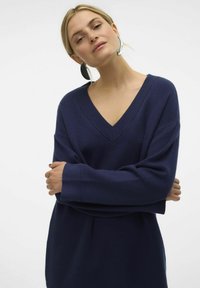 Marineblaues übergroßes Sweatshirt mit tiefem V-Ausschnitt, gerippten Bündchen und strukturiertem Finish. Das Modell posiert mit verschränkten Armen und trägt große silberne Ohrringe.