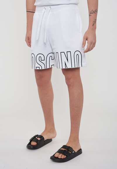 Shorts bianchi in misto cotone, con grande logo nero "MOSCHINO" all'orlo. Abbinati a sandali neri con doppia fibbia.