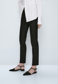 Svarta slim fit-byxor med en slät yta, kompletterade med en vit skjorta med knappar. Modellen bär svarta spetsiga ballerina-skor med ankelremmar.