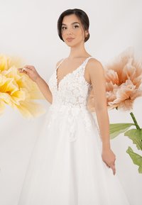 Ein weißes Tüllkleid mit Spitzenverzierungen am Oberteil, das einen tiefen V-Ausschnitt und dünne Träger hat, vor großen Papierblumen-Hintergründen.