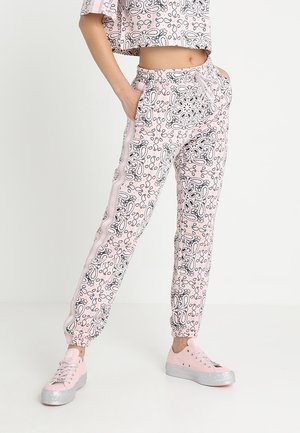Personne portant un pantalon de jogging rose à motif cachemire avec des bandes latérales et des baskets roses assorties à semelles argentées.