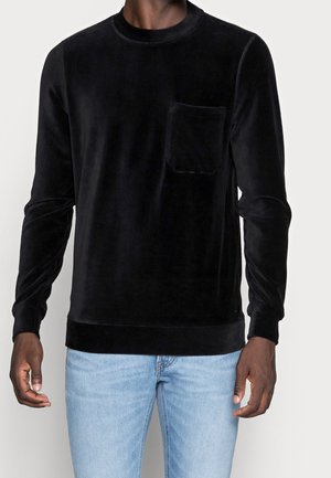 Mężczyzna w czarnej, welurowej koszuli z długim rękawem i kieszenią na piersi oraz jasnoniebieskich dżinsach, stojący na jednolitym tle.