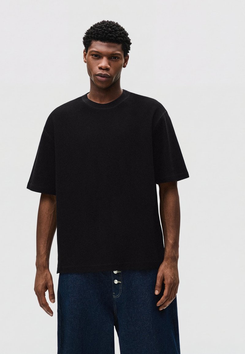 Jeune homme aux cheveux courts et bouclés portant un t-shirt noir uni oversized et un jean large bleu foncé sur fond clair.