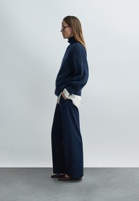 Pull à col roulé bleu marine, pantalon large à fines rayures, chemise blanche en dessous, et ballerines marron. Textures lisses et coupe décontractée.