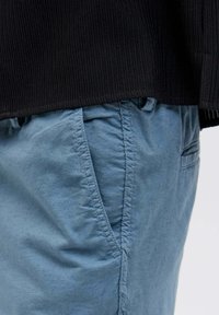 Maglione nero a coste con orlo sopra pantaloni leggeri blu, texturizzati, con tasche visibili e dettagli di cucitura distintivi ai lati.