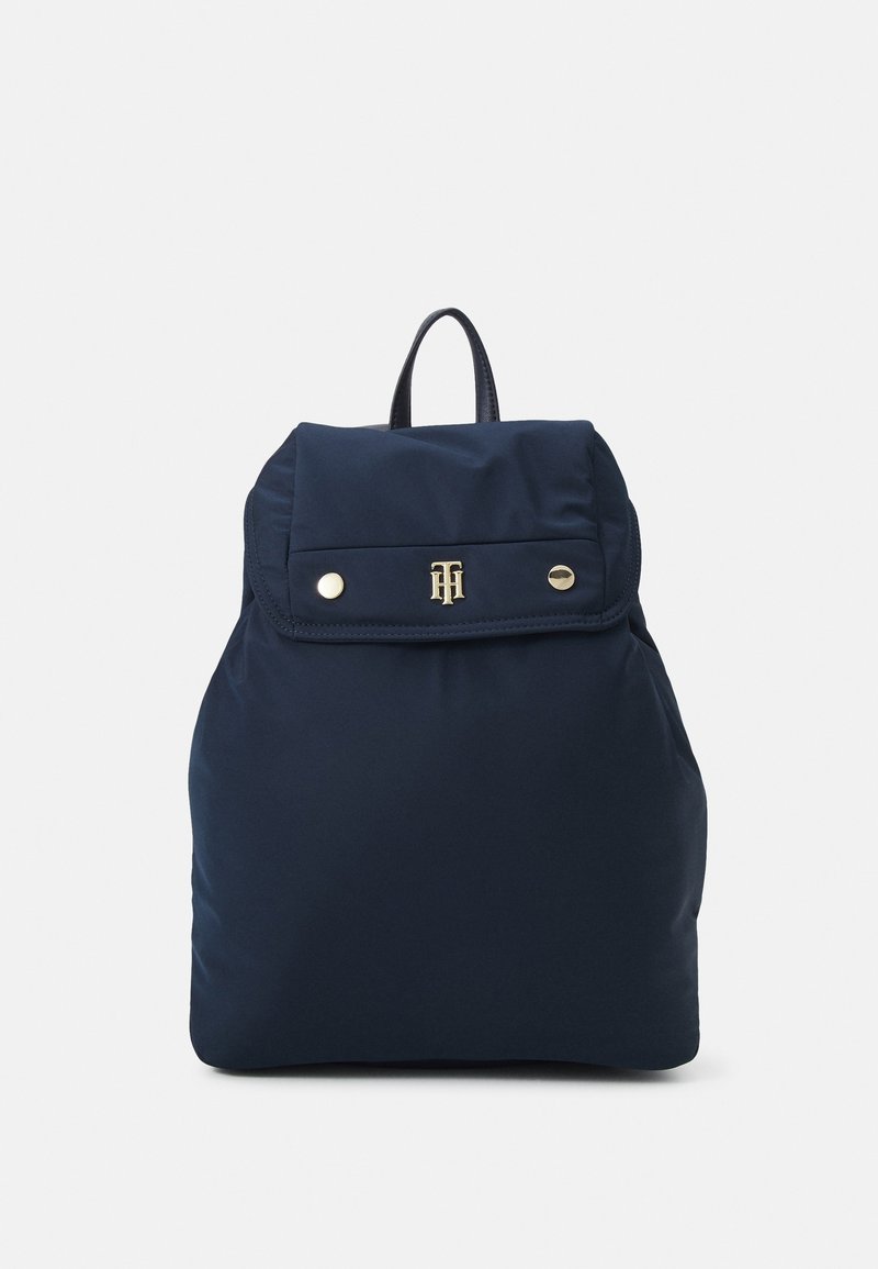 Mörkblå ryggsäck med toppflik, två gyllene tryckknappar, en gyllene monogramlogotyp och ett svart bärhandtag på vit bakgrund.