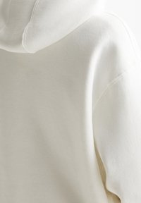 Close-up van een off-white zachte stoffen hoodie die gedetailleerde stiksels op de schouder en de naad van de capuchon laat zien.