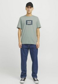 Zeleni pamučni majica s kvadratnim plavim logotipom na kojem piše "JACK & JONES." Uparena s širokim plavim trapericama i crnim tenisicama.