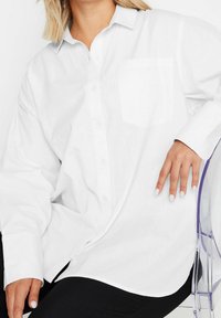 Woman wearing loose white button-up shirt with chest pocket, seated on transparent chair, with black pants visible.