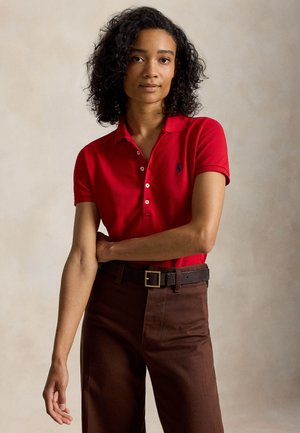 Γυναίκα με σγουρά μαλλιά που φοράει κόκκινο πόλο με κοντά μανίκια και καφέ παντελόνι, στέκεται με το ένα χέρι ακουμπισμένο στη μέση της.