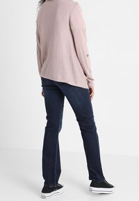 Hellrosa langärmliges Hemd, dunkelblaue Jeans mit geradem Bein und schwarze Turnschuhe mit weißen Sohlen, Rückansicht.