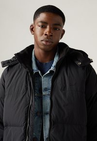 Black puffer jacket with a zip closure, worn over a denim shirt. The jacket features a hood and a textured finish.