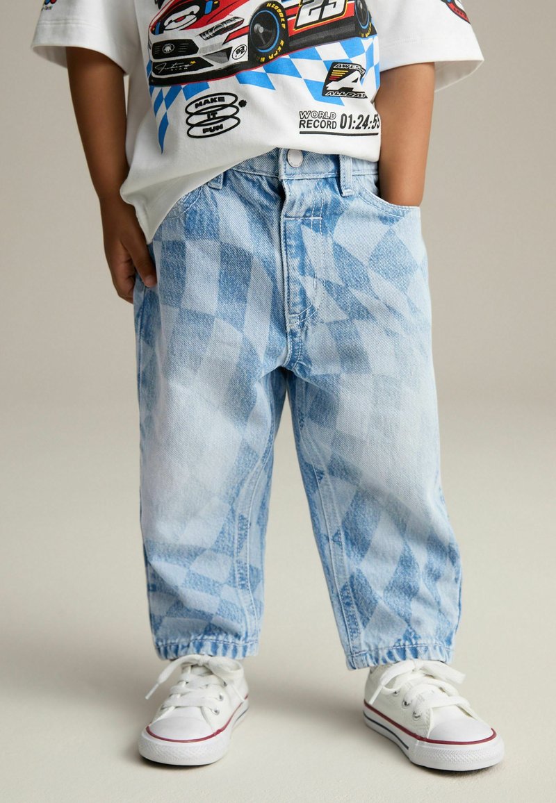Kind trägt hellblaue karierte Jeans, weiße Sneakers und ein weißes T-Shirt mit Rennsportdesign und Auto-Grafik.