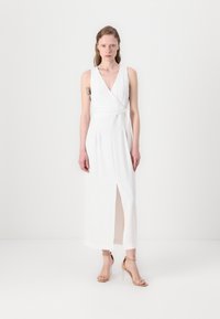 Rochie albă cu decolteu adânc în V, slit lateral și design pliat, asociată cu sandale nude cu curea. Material neted și siluetă până la podea.