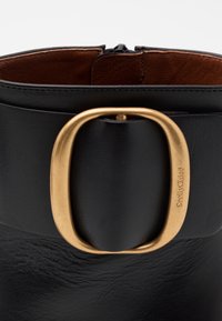 Černá kožená kotníková bota s kulatou zlatou sponou a hnědým vnitřním podšívkou. Zapínání na zip na zadní straně pro snadné obutí.