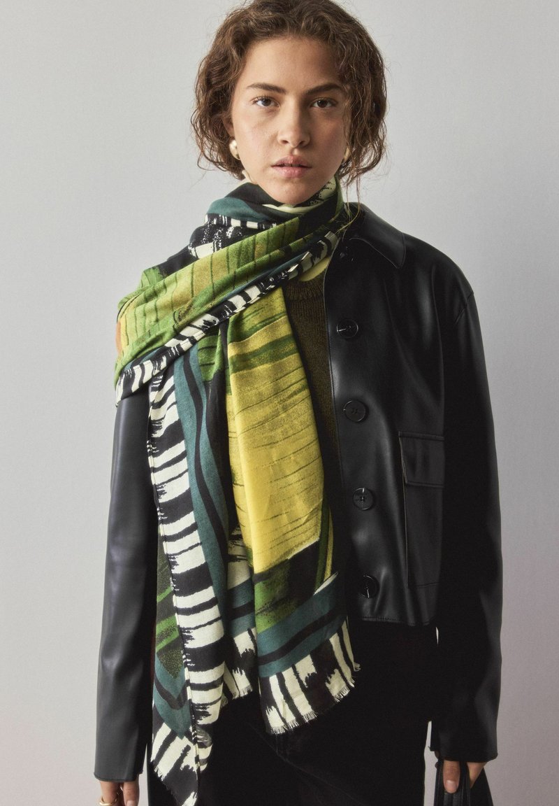 Daugiaspalvė šalikėlis su žaliais, geltonais ir juodais raštais; pagamintas iš minkštos medžiagos; užsegtas ant juodos odinės striukės su sagomis.