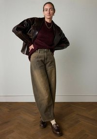 Bruine leren jas, bordeauxrode trui, olijfgroene wijde jeans en donkerbruine schoenen met een gesp. Gezet op een visgraat houten vloer.