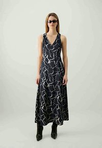 Fekete mintás maxiruha V-kivágással és ujjatlan kialakítással. Absztrakt fehér formákat tartalmaz, és bokáig ér. Fekete csizmával viselve.