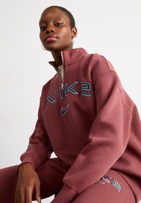 Sweatshirt com fecho de correr, de cor rosa escuro, com gola alta, apresentando o logotipo da Nike em azul e cinza no peito. Tecido liso e macio.