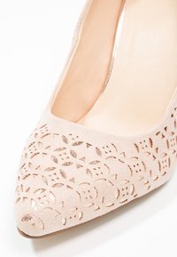 Chaussure plate beige à bout pointu avec motif floral découpé laissant apparaître une couche métallique dorée en dessous.