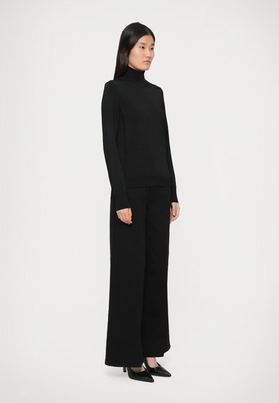 Kvinde iført en sort turtleneck sweater, brede sorte bukser og sorte spidse hæle, stående mod en ensfarvet hvid baggrund.