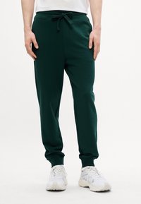 Pantaloni jogger verdi in tessuto morbido con waistband elastico, laccetto, tasche laterali e polsini a costine. Indossati con scarpe da ginnastica bianche.