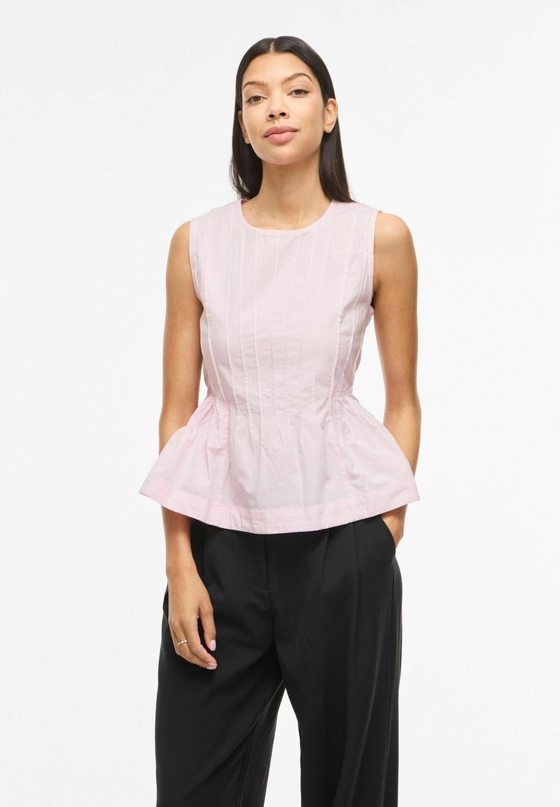 Femme aux longs cheveux foncés portant un haut péplum rose pâle sans manches et un pantalon noir ample, debout avec une main dans la poche.