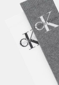 Dos pares de calcetines de algodón; uno gris con un logo negro prominente, el otro blanco con un logo tenue; ambos presentan una textura suave.