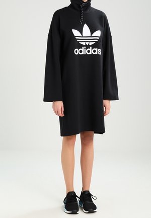 Persoon draagt een zwarte Adidas hoodie-jurk met lange mouwen en een hoge kraag, gecombineerd met zwarte sportschoenen met blauwe accenten.