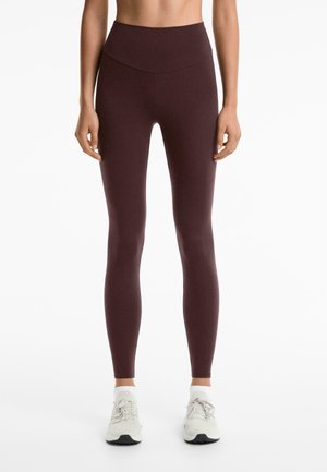 Vrouw in hooggemeten, donkerbruine leggings en witte sneakers, staande met ontspannen armen langs haar zij tegen een witte achtergrond.