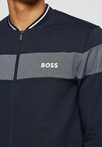 Marinfärgad zip-up-jacka med ribbad krage, horisontell grå rand och broderad "BOSS"-logotyp i vitt på bröstet. Slät tygtextur.