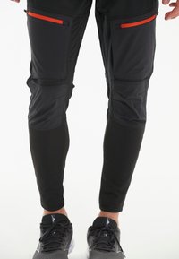 Schwarze Sporthose mit roten Reißverschlusstaschen, ausgestattet mit strukturierten Einsätzen und einem enganliegenden Design. Getragen mit schwarzen Sportschuhen.