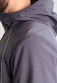 Homme portant une veste imperméable gris foncé avec une capuche et une couture de fermeture éclair jaune visible au niveau de l'épaule.