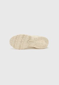 Beige treningssko såle med teksturert mønster, med spor for grep og en synlig merkevare-logo. Glatt gummimateriale.