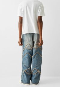 Unisex white T-shirt paired with loose blue denim jeans featuring diamond-shaped pockets with frayed edges and a textured finish.