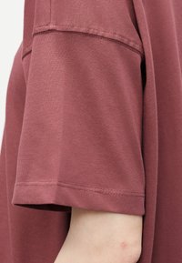 Gros plan sur le bras d'une personne portant une chemise mauve ample à manches courtes avec des coutures visibles et une partie d'une main pâle sous la manche.