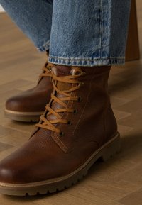 Bottines en cuir marron avec une finition texturée, bout rond et lacets beige. Elles présentent une semelle robuste et des détails de couture.