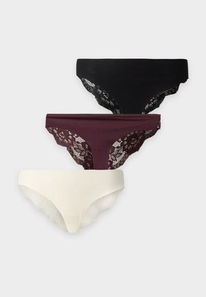 Donna in piedi a piedi nudi contro uno sfondo semplice, indossa un reggiseno marrone e biancheria intima bianca, guarda leggermente di lato con un braccio incrociato.