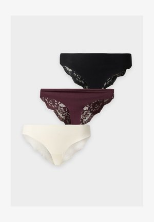 Donna in piedi a piedi nudi contro uno sfondo semplice, indossa un reggiseno marrone e biancheria intima bianca, guarda leggermente di lato con un braccio incrociato.