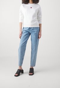 Donna che indossa una felpa bianca a maniche lunghe, jeans alta vita azzurri chiari e mules con tacco aperto neri, in piedi davanti a uno sfondo semplice.