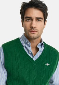 Groene kabelgebreide vest van wol, met een V-hals ontwerp. Draag het over een lichtblauw gestreept shirt met een button-down kraag.
