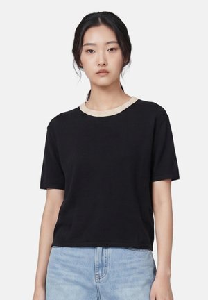 Junge Frau mit zurückgebundenen dunklen Haaren, die ein schwarzes kurzärmeliges Shirt mit beigem Kragen und hellblaue Jeans vor weißem Hintergrund trägt.