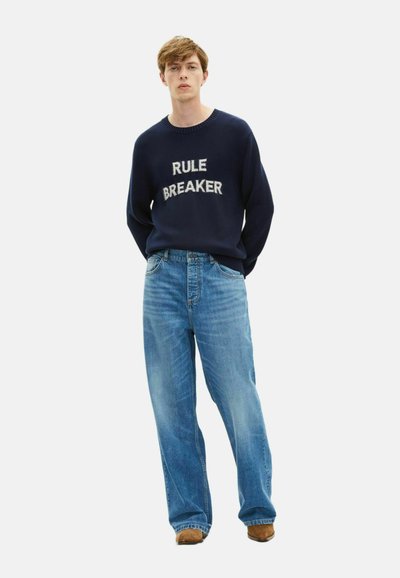 Maglione blu navy con il testo bianco "RULE BREAKER", abbinato a jeans in denim chiaro a gamba larga e scarpe marroni, su uno sfondo semplice.