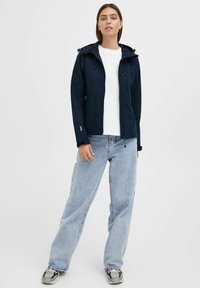 Mørkeblå soft-shell jakke med hette, glidelås foran og sidelommer, kombinert med løse lyseblå jeans og svart-hvite sneakers.