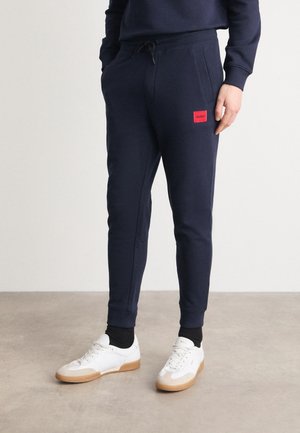 Personne portant un jogging bleu marine avec un patch logo Hugo rouge, des chaussettes noires et des baskets blanches, debout sur un sol en béton.