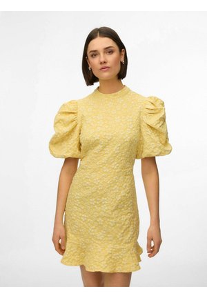 Donna che indossa un vestito giallo a motivo floreale con maniche arricciate e orlo a volant, in piedi davanti a uno sfondo bianco uniforme.